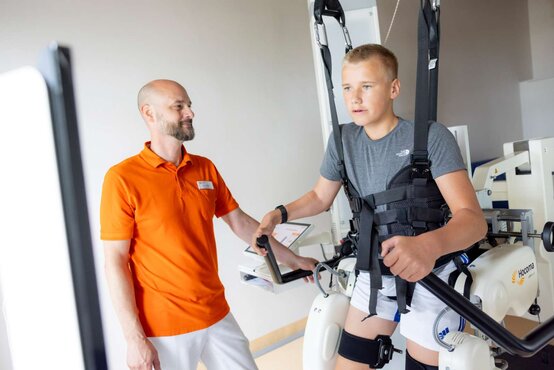 Physiotherapeut im orangefarbenen Poloshirt betreut Jungen im Lokomat-Gangtrainer, gesichert mit Gurten, heller Therapieraum