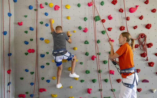 Kind klettert gesichert an bunter Kletterwand; Therapeutin im orangefarbenen Shirt sichert mit Seil im Bewegungsraum