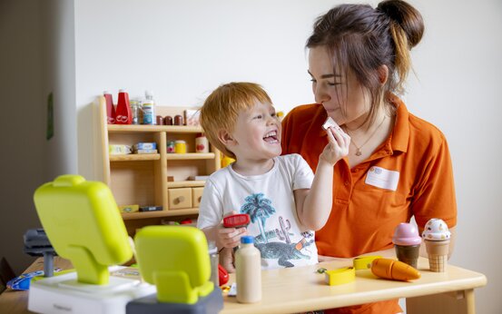 Junge mit roten Haaren spielt lachend mit erwachsener Frau in orangefarbener Kleidung an Holztisch, Spielzeugeis, Spielzeugmöhre und Registrierkasse