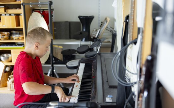 Junge spielt am Digitalpiano mit farbigen Markierungen, Gong, Gitarren und Schlagzeug im hellen Musiktherapieraum im Hintergrund