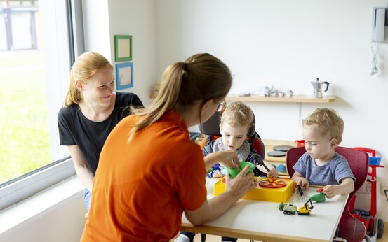 Zwei kleine Kinder spielen am Tisch mit Lernspielzeug, Therapeutin im orangefarbenen Oberteil hilft, zweite Frau sitzt entspannt am Fenster