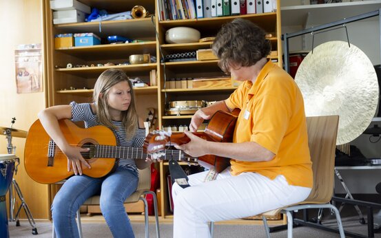 Mädchen mit Gitarre erhält Gitarrenstunde von Musiktherapeutin im orangefarbenen Poloshirt, beide sitzen im Musikraum