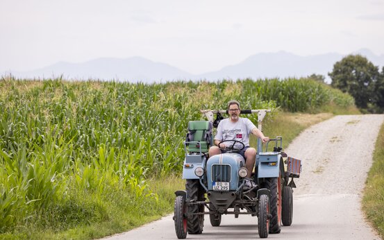 Mann mit Brille fährt bei Sonnenschein auf altem blauen Traktor einen Feldweg, neben ihm ein Kindersitz
