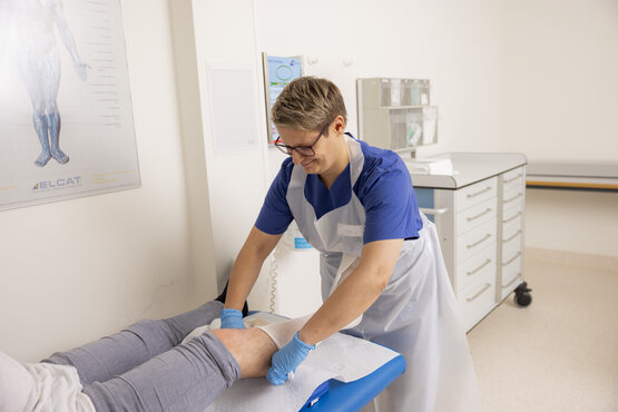 Medizinisches Fachpersonal in blauer Dienstkleidung legt einem Patienten auf der Liege einen Verband am Unterschenkel an