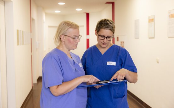 Zwei Personen in blauer medizinischer Kleidung betrachten gemeinsam ein Klemmbrett im hellen Flur, beide mit Namensschild und Brille