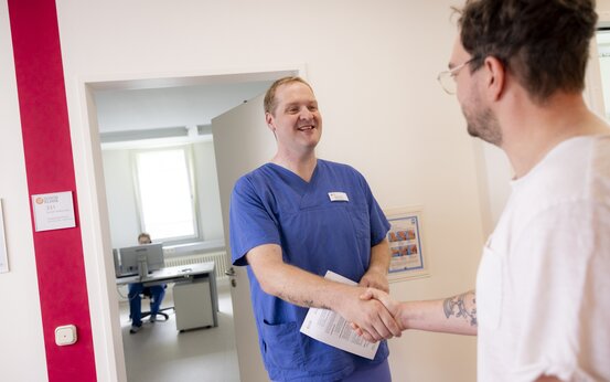 Zwei Männer geben sich im hellen Klinikflur die Hand, einer in blauer medizinischer Kleidung mit Papier, einer im weißen T-Shirt mit Brille