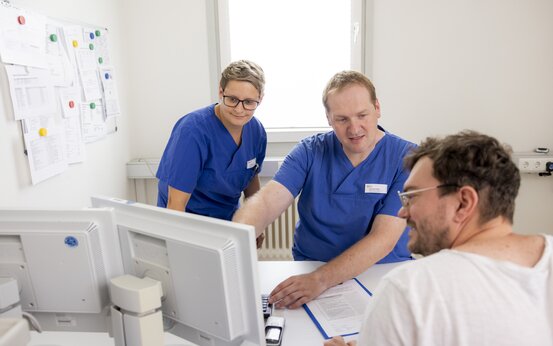 Zwei medizinische Fachkräfte in blauer Arbeitskleidung sitzen am Computer und besprechen mit einem Mann im weißen Shirt Unterlagen
