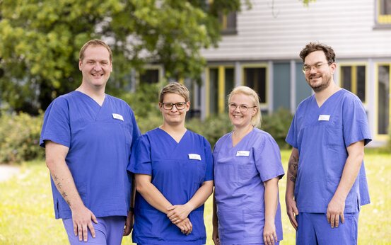 Vier Personen in blauer Arbeitskleidung mit Namensschild stehen lächelnd auf einer Wiese vor Bäumen und Klinik