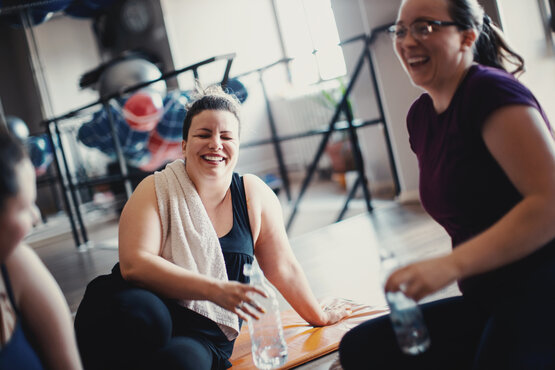 Drei Frauen machen lachend Pause beim Training im Fitnessstudio, halten Wasserflaschen in der Hand