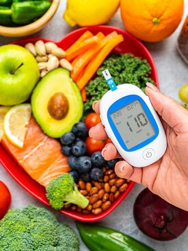 Hand hält Blutzuckermessgerät mit 110 mg/dl, herzförmige Schale mit Avocado, Lachs, Apfel, Karotten, Nüssen und Gemüse