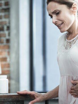 Frau in heller Kleidung steht in moderner Küche, hält sich mit schmerzverzerrtem Gesicht den Bauch, Wasserflasche und Medikamente auf Arbeitsplatte