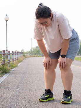 Übergewichtige Person stützt sich erschöpft beim Sport auf einer ruhigen Straße, trägt Smartwatch.