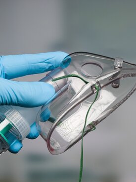 Hand mit blauem Handschuh hält Sauerstoffmaske mit grünem Band und Schlauchanschluss. 