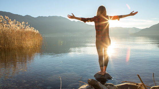 Person steht barfuß auf Stein am klaren See vor Bergkulisse, ausgebreitete Arme