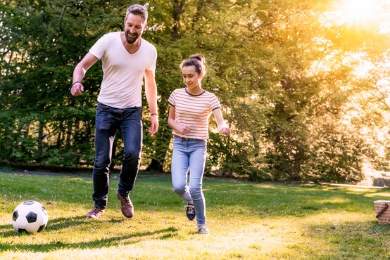 Vater und Tochter spielen gemeinsam Fußball auf sonniger Wiese im Park, beide bewegen sich aktiv