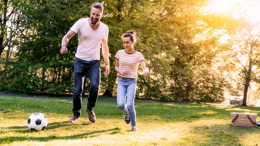 Vater und Tochter spielen gemeinsam Fußball auf sonniger Wiese im Park, beide bewegen sich aktiv