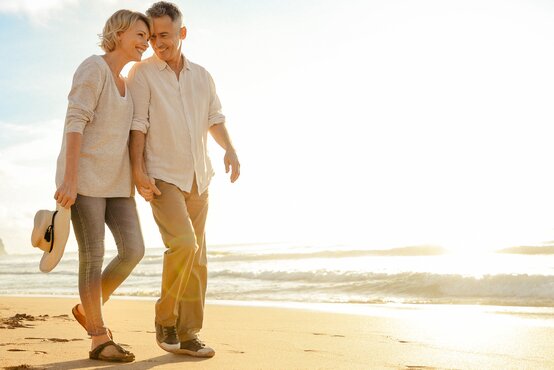 Glückliches älteres Paar spaziert barfuß und sicher am sonnigen Strand, hält sich an den Händen