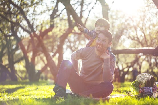 Vater sitzt entspannt mit Kind auf dem Schoß auf Picknickdecke im grünen Park, Picknickkorb mit Obst