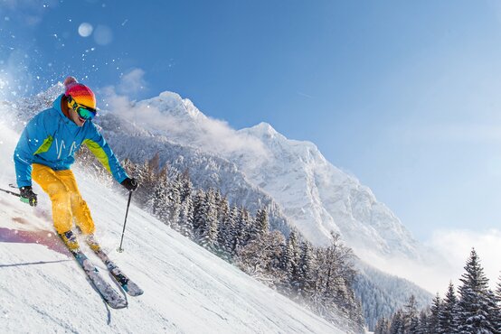 Person in gelber Hose und blauer Jacke fährt auf Ski mit Skistöcken auf schneebedeckter Piste im Gebirge