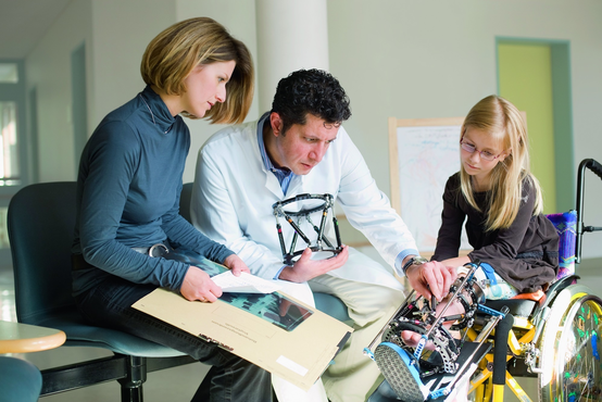 Art erklärt Frau und Mädchen nach OP die Fixateur des Mädchens, sitztz im Rollstuhl