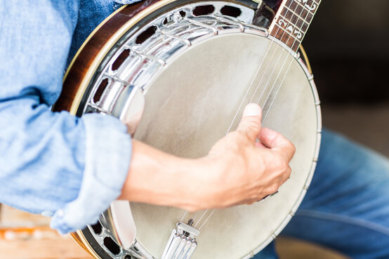 Erwachsene Person in Jeans sitzt auf Stuhl und spielt Banjo, Fokus auf rechte Hand beim Saitenzupfen