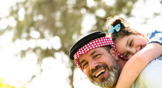 Lachender arabischer Vater mit rot-weißer Kufiya, hält Kind mit blauer Schleife auf den Rücken