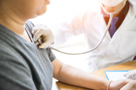 Arzt mit Mundschutz und Handschuhen untersucht erwachsenen Patienten mit Stethoskop am Brustkorb in heller Klinik