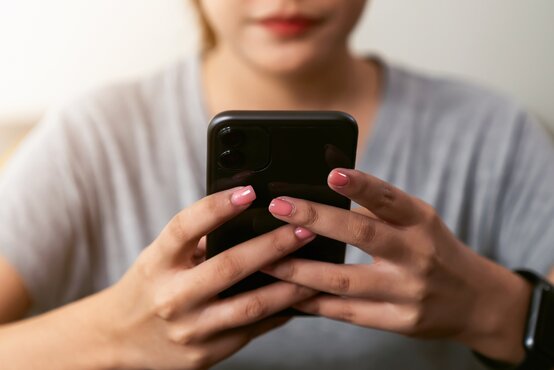 Junge Frau im grauen T-Shirt hält mit beiden Händen ein schwarzes Smartphone, Hände mit rosa lackierten Fingernägeln und Smartwatch