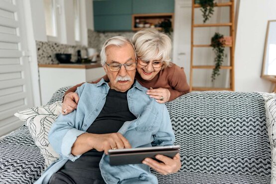 Älterer Mann mit Tablet sitzt auf Sofa, ältere Frau umarmt ihn von hinten, beide tragen Brille, moderne Küche im Hintergrund