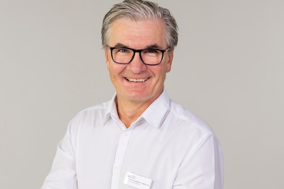 Mann mit grauen, nach hinten gekämmten Haaren und schwarzer Brille lächelt freundlich in die Kamera.