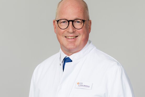 Stefan Middeldorf. Porträt eines männlichen Arztes mit Brille und Glatze im weißen Kittel, blaues Hemd, Krawatte