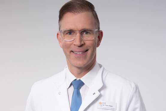 Prof. Dr. Thilo Patzer. Freundlich lächelnder Arzt mit Brille und Namensschild im weißen Kittel