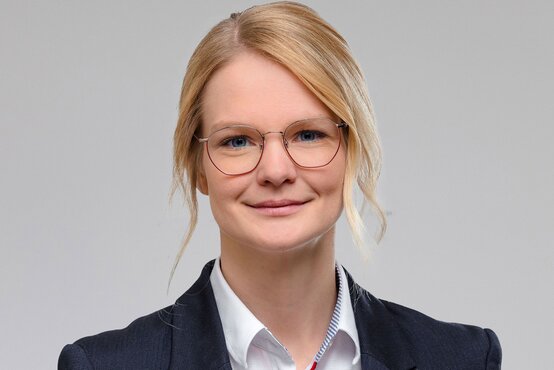 Porträt von Julia Tamborini mit blonden Haaren, runder Brille, weißem Hemd und dunklem Sakko vor hellem Hintergrund