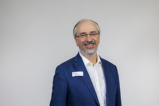 Porträt von Prof. Dr. Christian Andreas Hillert, Mann mit grauem Haar, Bart und Brille im blauen Sakko vor hellgrauem Hintergrund