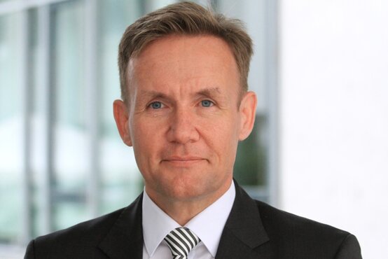 Porträt eines Mannes mit kurzem braunem Haar, weißem Hemd, gestreifter Krawatte und schwarzem Anzug vor Glasfassade