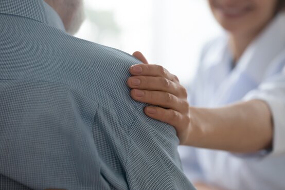 Pflegekraft legt unterstützend die Hand auf die Schulter eines älteren Mannes