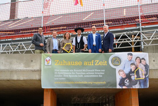 Sechs Erwachsene stehen auf Baustelle vor Gerüst und Banner „Zuhause auf Zeit“ zum Ronald McDonald Haus, eine Frau hält gelbes Hausrahmen-Element