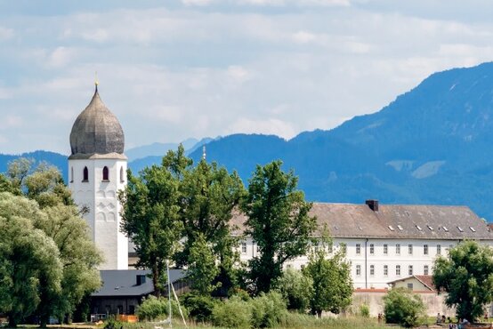 Großes weißes Gebäude mit vielen Fenstern direkt am Ufer eines Sees, links Kirche mit Zwiebelhaube, dahinter bewaldete Alpen und blauer Himmel