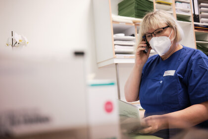 Medizinische Fachkraft mit blonden Haaren, Brille und FFP2-Maske telefoniert im und prüft medizinische Dokumente
