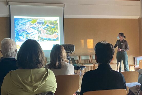 Präsentation in Klinik: Referentin mit Maske und Namensschild zeigt Lageplan des Klinikgeländes auf Leinwand vor sitzenden Teilnehmenden