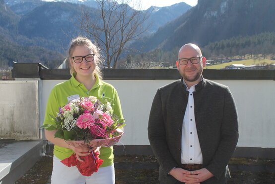 Junge Frau im grünen Poloshirt mit Blumenstrauß und Mann im weißen Hemd vor Alpenpanorama
