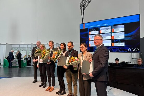 Sechs Personen auf Bühne bei DGP-Preisverleihung, vier mit Blumensträußen und Urkunden, Präsentationsfolie im Hintergrund