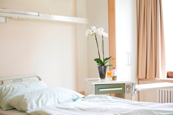 Helles, gepflegtes Klinik-Patientenzimmer mit Krankenbett, Nachttisch, Orchidee, Glas Orangensaft und Fenster mit Tageslicht