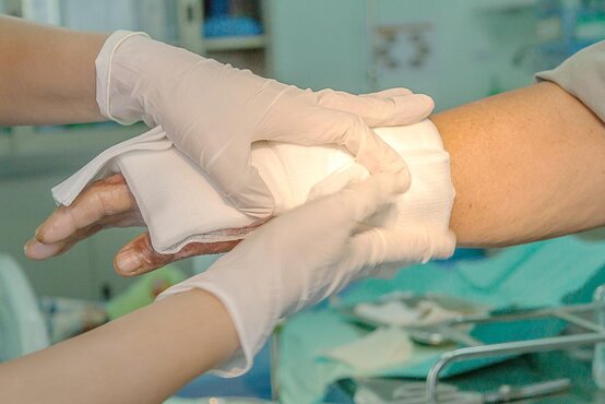 Zwei behandschuhte Hände wickeln sterilen Verband um verletzte Hand im klinischen Umfeld, Instrumente und blaues Tuch im Hintergrund