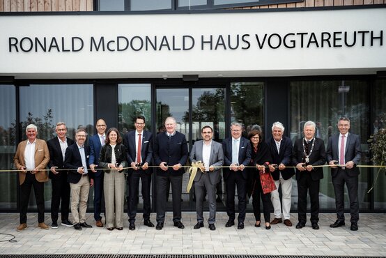 Feierliche Banddurchschneide-Zeremonie vor dem Ronald McDonald Haus Vogtareuth: Fünfzehn festlich gekleidete Erwachsene halten gelbes Band