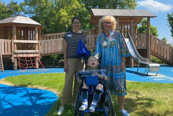 Zwei Frauen und ein Kind im spezialisierten blauen Rollstuhl vor barrierefreiem Holzspielplatz bei Sonnenschein