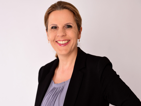 Porträtfoto von Astrid Angele, Frau mit schulterlangem, blondem Haar, Brille und dunklem Oberteil vor hellem Hintergrund