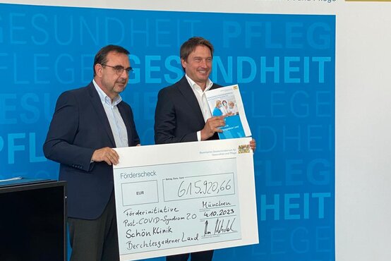 Zwei Männer im Anzug übergeben symbolischen Förderscheck für Post-COVID-Syndrom an die Schön Klinik Berchtesgadener Land