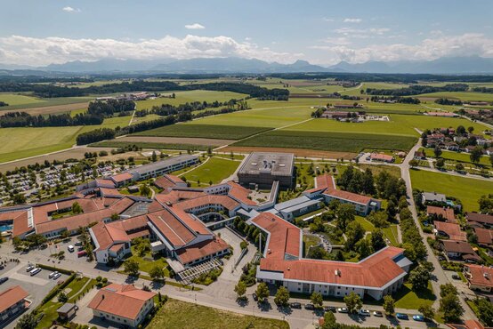 Luftaufnahme der Schön Klinik Vogtareuth mit modernen Gebäuden, roten Dächern, Wegen, Parkplätzen und umliegender Natur mit Bergen im Hintergrund