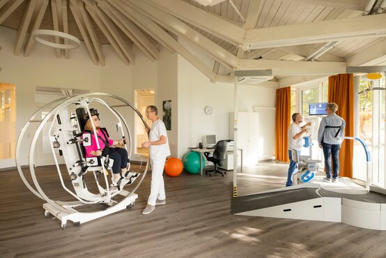 Therapieraum mit großem weißen Reha-Gerät, Patientin mit Gurten gesichert, Plattform mit Rampe, Monitor und zwei Gymnastikbällen